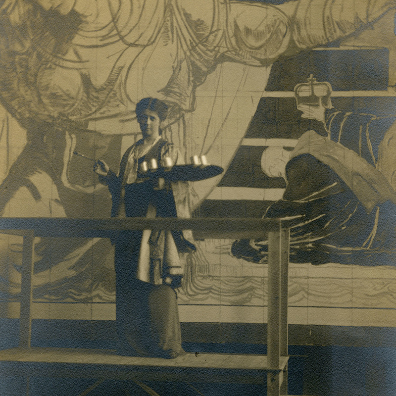 Full-length portrait of Oakley, attired in a smock and dress, and standing on a platform and holding a paint palette in front of a large painted figure of a woman with outstretched arms.
